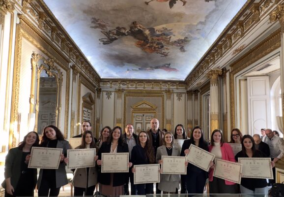 Foto di gruppo della cerimonia conclusiva del Master GMC 2024, con i partecipanti che mostrano i loro diplomi in una sala ornata da decorazioni dorate e affreschi sul soffitto.